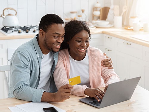 young adult couple seated at kitchen table using a laptop and holding a credit card